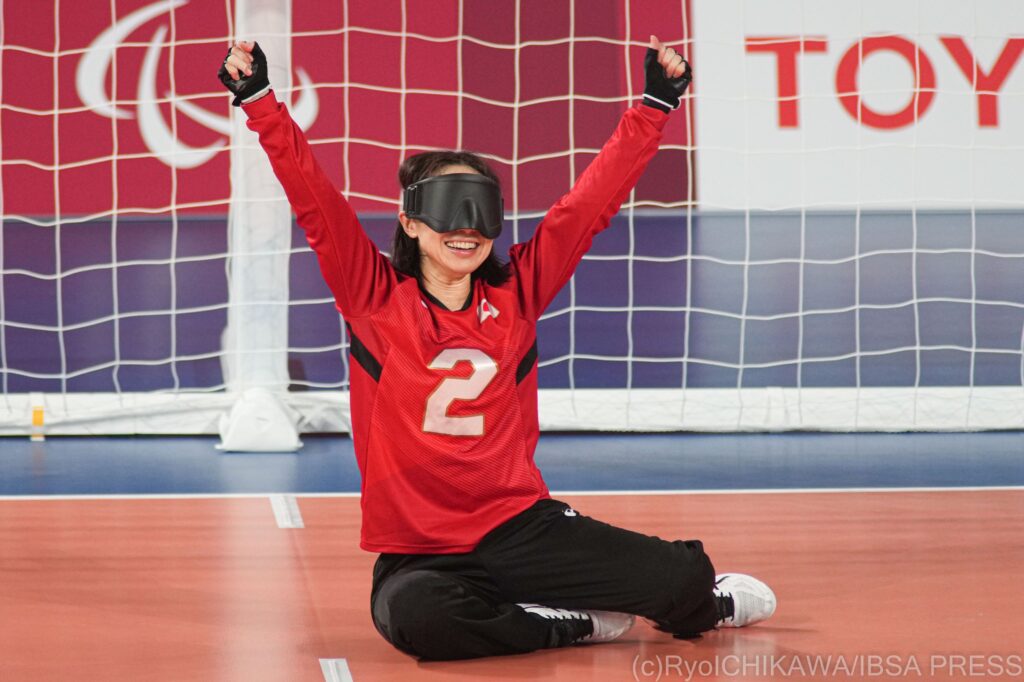 A female goalball player from Japan sits in goal and raises her arms in the air with delight