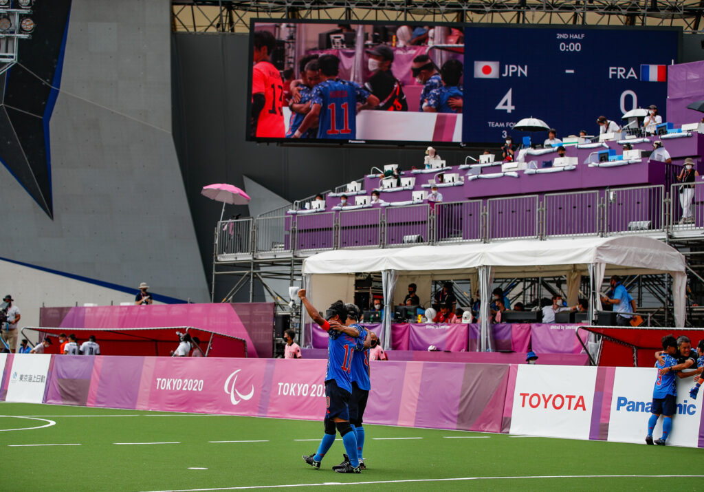 Two Japanese players embrace as a wide shot of the blind football venue is shown